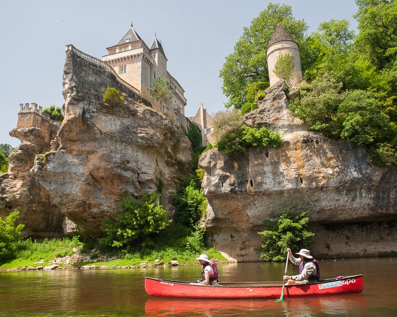 A red canoe glides on calm waters near steep, rocky cliffs. Towering structures, including a castle and a turret, are nestled among lush greenery. The landscape is complemented by the contrasting textures of stone and trees, offering a scenic view of the surrounding area.