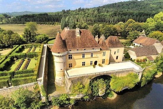 An aerial view showcases a charming stone villa set on the bank of a river, surrounded by lush greenery. The structure features a distinctive turret, large windows, and a terrace overlooking landscaped gardens, all contributing to a peaceful natural setting.