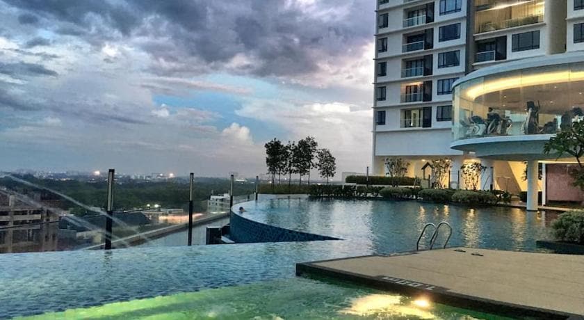 A modern swimming pool area showcases a large infinity edge that overlooks panoramic views. Lush greenery is visible along the edge, while the surrounding building features contemporary architecture with multiple windows. A cloudy sky creates a dramatic backdrop for this serene space.