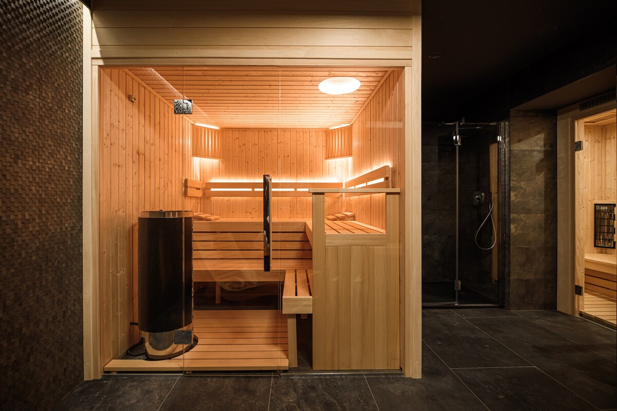 A modern sauna room features wooden benches and walls, illuminated by soft ambient lighting. A glass door provides visibility, while a wood-burning stove is positioned against one wall. The floor is surfaced with dark tiles, adding to the serene ambiance.