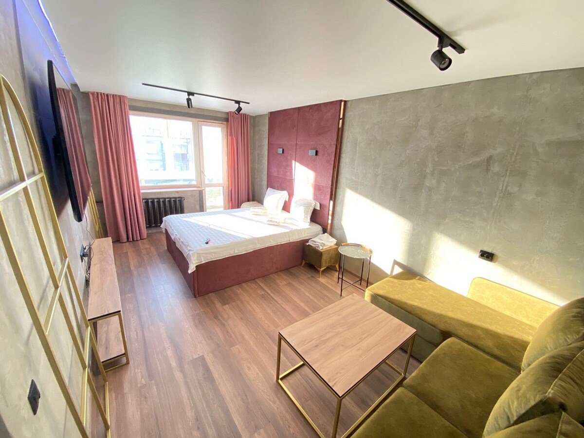 A spacious room features a comfortable sleeping area with two beds arranged neatly. Light streams through a large window adorned with soft pink curtains. A cozy seating area is provided with a green sofa and a coffee table, complementing the warm wooden flooring.