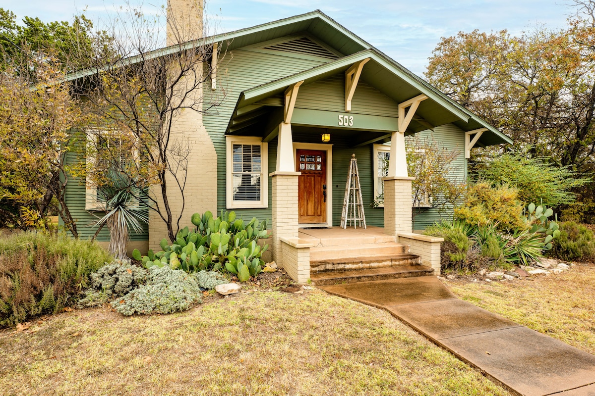 The exterior of the historic craftsman home displays a green facade, with a welcoming porch and wooden front door. Lush greenery surrounds the entrance, enhancing the inviting atmosphere, while rocky landscaping offers character. A pathway leads to the door, flanked by low shrubs and desert plants.