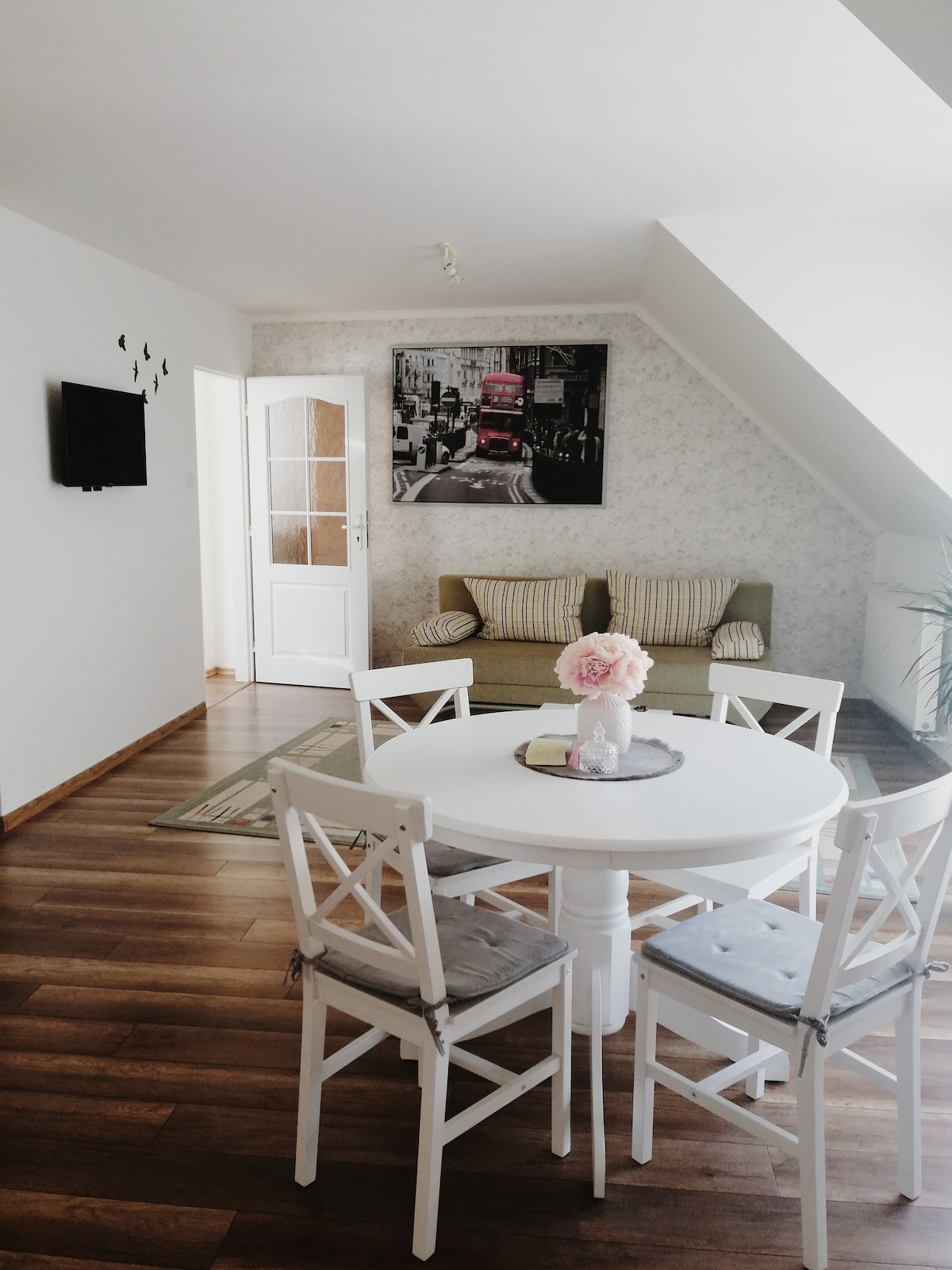 A cozy dining area features a round white table surrounded by four chairs with soft cushions. A green sofa is positioned along the wall, with a framed urban print above it. Natural light illuminates the polished wooden floor, enhancing the inviting ambiance.