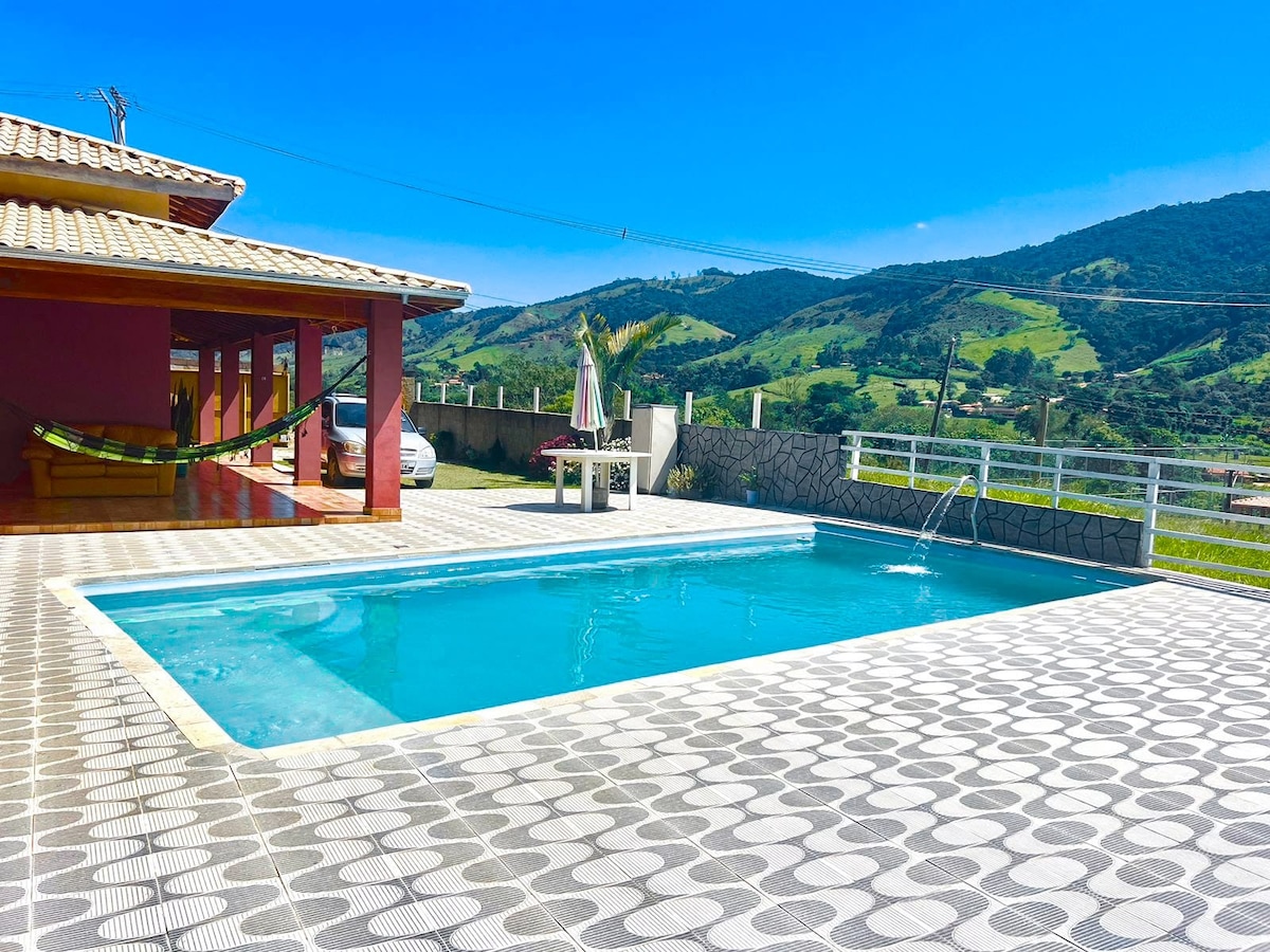 A spacious outdoor area is highlighted by a rectangular swimming pool surrounded by a patterned terrace. The view includes green hills in the background under a clear blue sky. A hammock is visible next to the pool, adding to the relaxing ambiance.