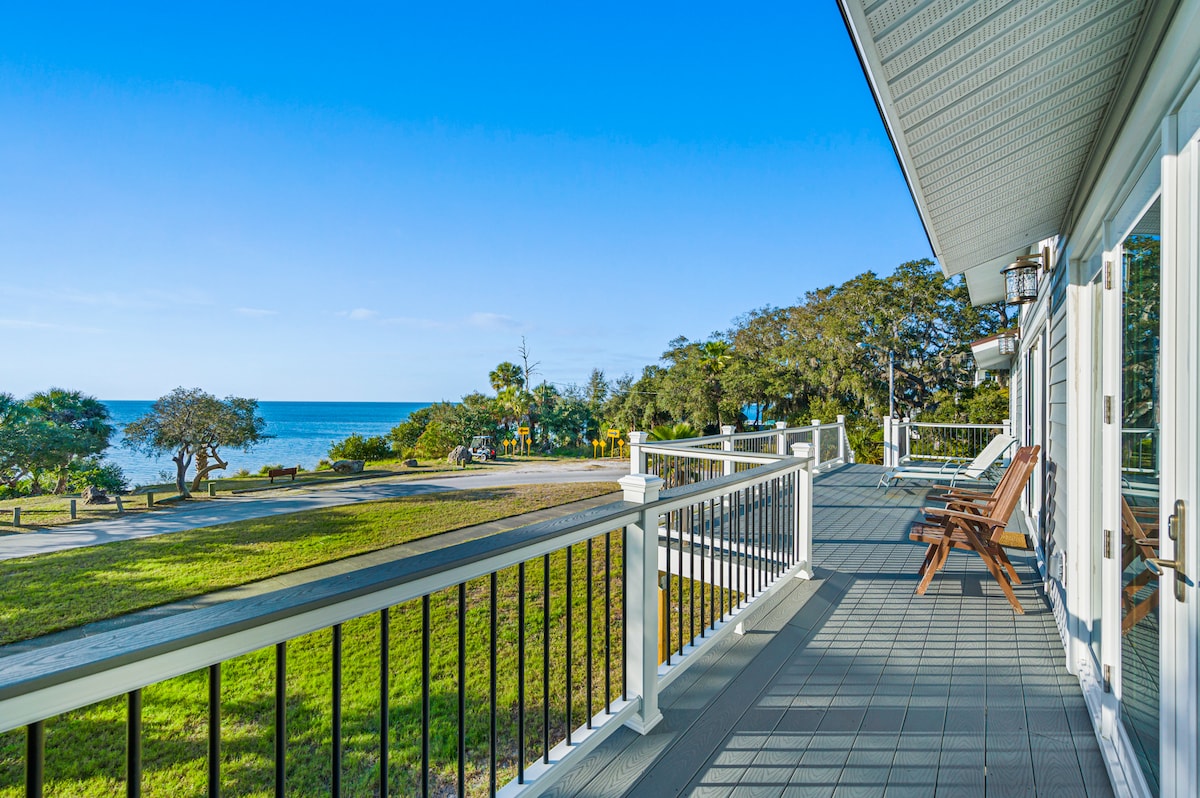 A spacious balcony is featured with comfortable wooden chairs, inviting relaxation. The view reveals a clear blue sky merging with the calm ocean, complemented by lush greenery in the foreground. The area offers a sense of tranquility, perfect for enjoying peaceful moments outdoors.