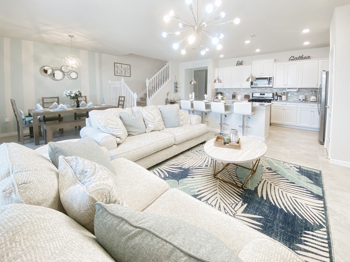 The open-concept living area features a spacious seating arrangement with light-toned sofas, complemented by a round coffee table. In the background, a dining table seating eight and a modern kitchen with white cabinetry and stainless-steel appliances are visible, illuminated by a stylish chandelier.