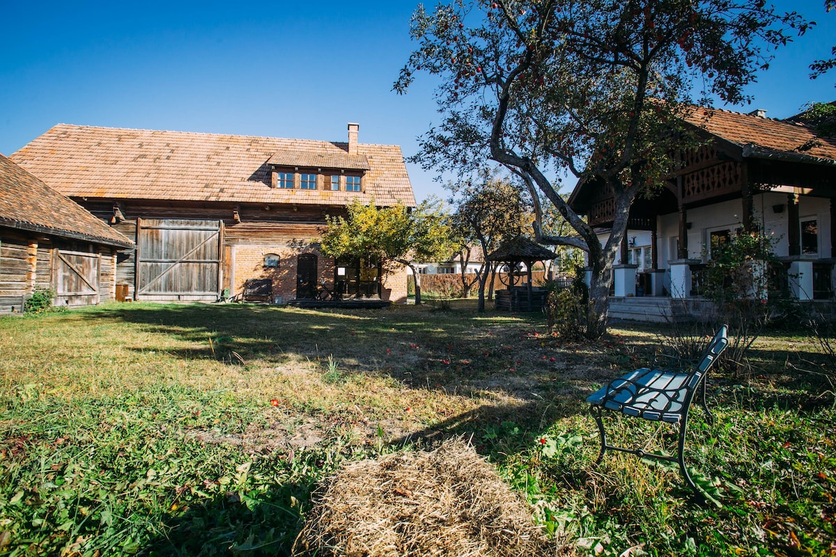 A sprawling garden area features a mix of grassy patches and scattered trees, with a rustic barn and traditional wooden house in the background. A single metal bench provides seating, offering a peaceful view of the surrounding landscape under a clear blue sky.
