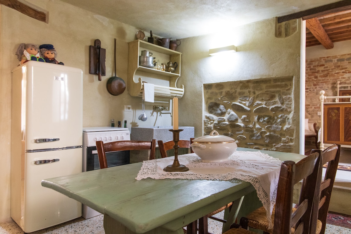 A rustic kitchen features a green wooden dining table surrounded by wooden chairs. An antique stove and modern fridge are visible, along with a white dish set atop a lace tablecloth. Natural stone accents and warm lighting enhance the inviting atmosphere.