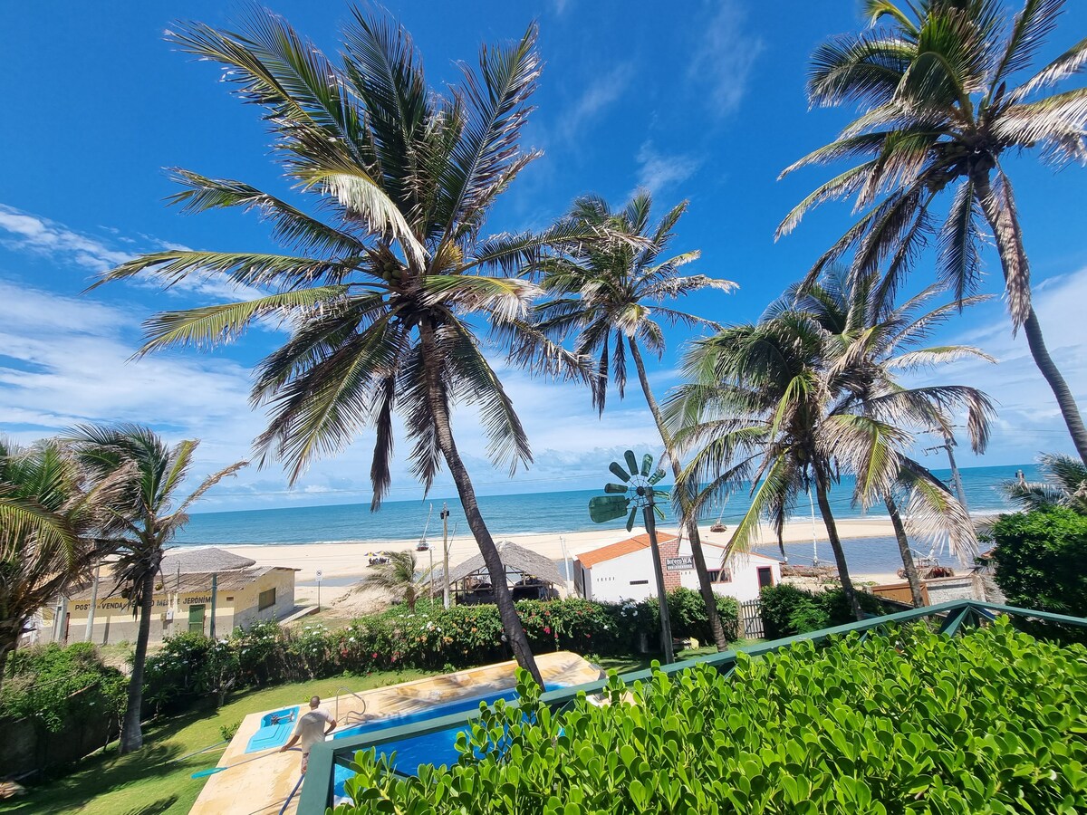 Tall palm trees sway gently in the breeze, framing a scenic view of the ocean. The expansive beach glistens under a clear blue sky, while a well-maintained pool and lush greenery enhance the inviting atmosphere of the outdoor space.