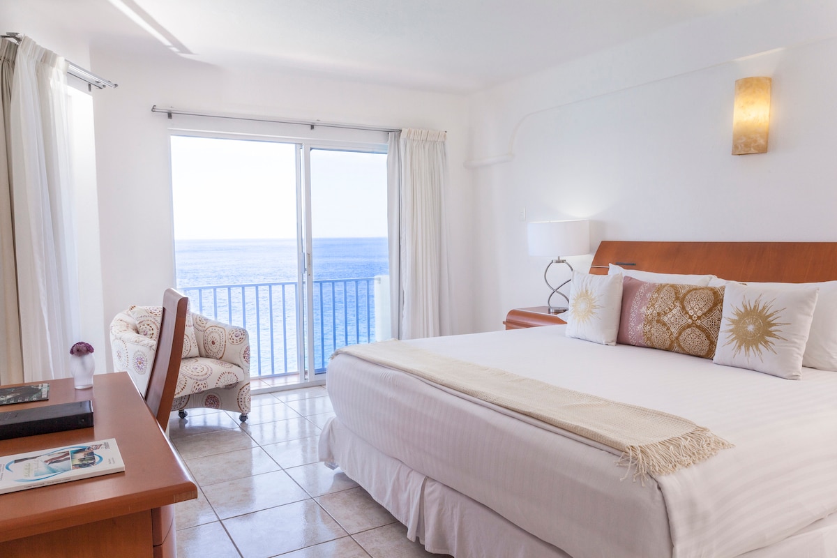 A bright and airy guest room features a spacious bed adorned with decorative pillows. Large glass doors offer a view of the ocean, while a cozy chair sits nearby. Natural light illuminates the space, accented by soft white curtains and warm wall fixtures.