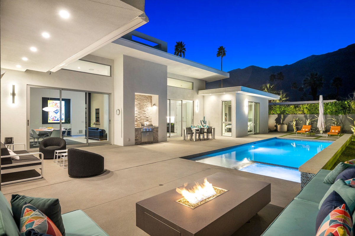 A contemporary outdoor area showcases a sparkling swimming pool surrounded by stylish furniture. A fire pit is positioned on one side, while a dining table is set nearby. The setting is illuminated by evening lights, with mountains visible in the background.