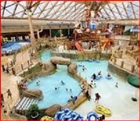 An expansive indoor water park is showcased, featuring a variety of water attractions, including slides and a lazy river. Families and children are seen enjoying the water amidst a vibrant tropical theme, with large structures and seating areas visible around the pool.