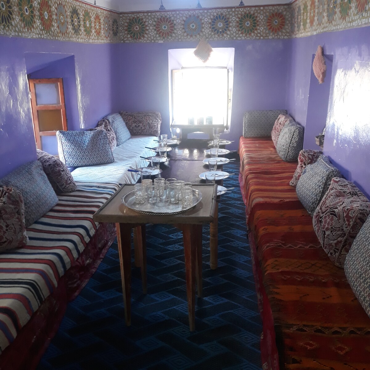 A vibrant sitting area showcases colorful textile seating arranged around a central table. Decorative patterns adorn the walls, and light filters through a window, accentuating the purple tones of the room. White tableware is neatly set on the table, inviting communal dining.
