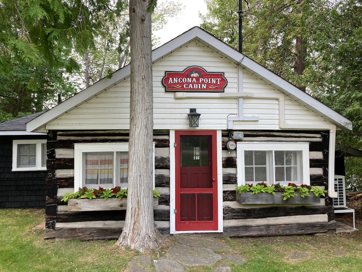 The exterior of a cozy log cabin features a welcoming red door and colorful flower boxes beneath the windows. The architecture reflects rustic charm, with wooden logs and a sloped roof. Surrounding greenery enhances the cabin's rustic appeal.