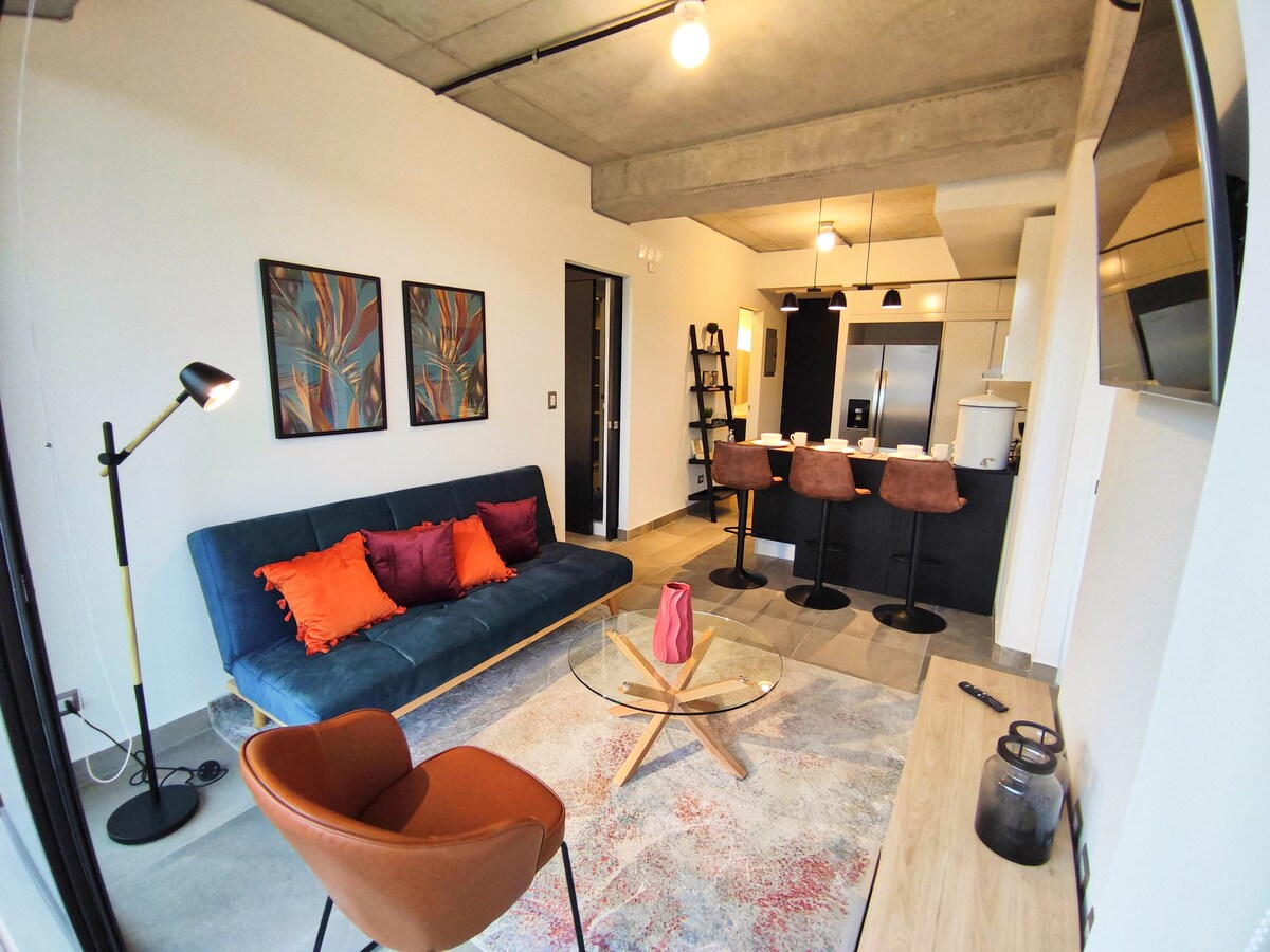 A modern living space features a blue sofa adorned with colorful cushions and a round table with a decorative vase. The kitchen is visible in the background, equipped with stainless steel appliances. A small dining area is set with stylish chairs, enhancing the overall contemporary design.