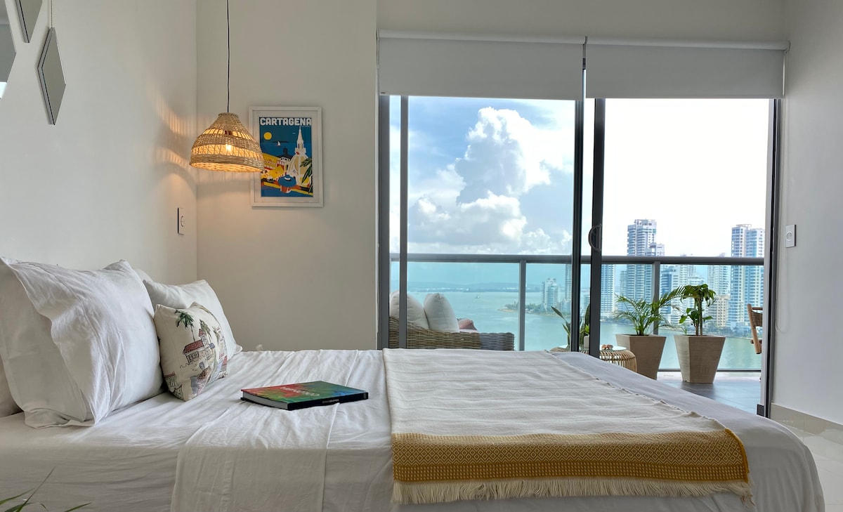 A spacious bedroom features a comfortable bed dressed in crisp white linens. Large glass doors provide access to a balcony with views of the bay and city. A ceiling lamp and wall art add a touch of style to the serene space.