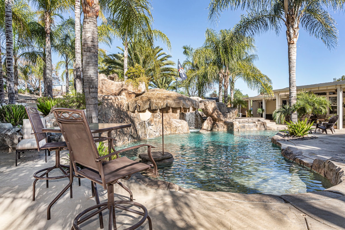 The backyard features a resort-style pool surrounded by palm trees and lush greenery. A rock waterfall is visible, enhancing the natural aesthetics. Several stylish lounge chairs are arranged nearby on a stone patio, providing comfortable seating for relaxation.