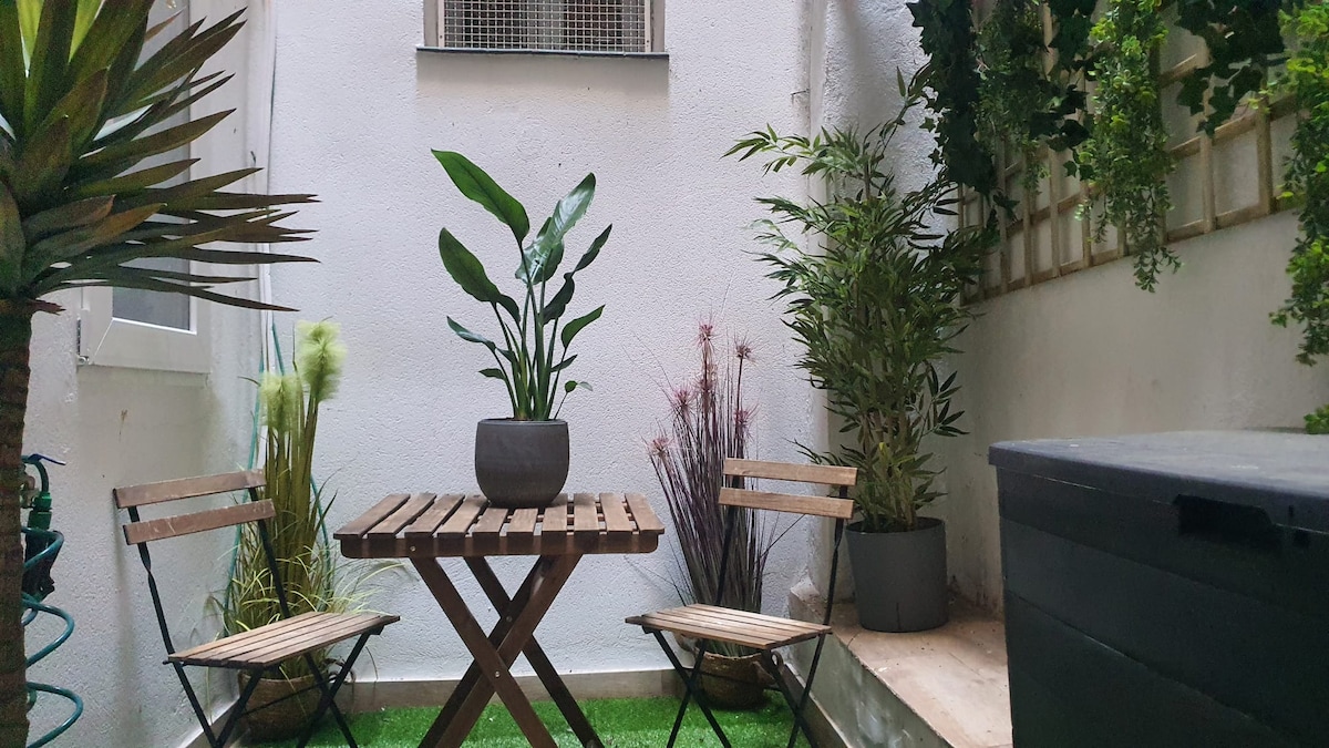 A small interior patio features a wooden table surrounded by two folding chairs. Lush green plants, including tall grasses and tropical foliage, provide a refreshing natural touch against the white walls. A storage box rests in one corner, enhancing the functionality of the space.