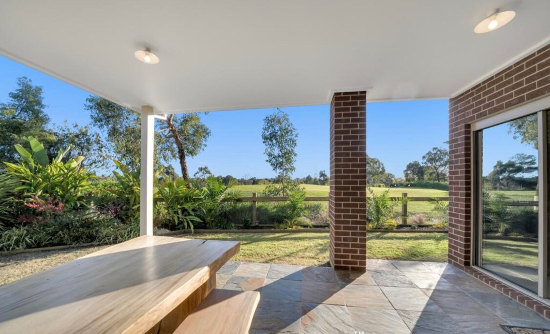 An outdoor alfresco area features a wooden table set against a backdrop of lush greenery and views of the nearby golf course. Sunlight streams through large windows, enhancing the inviting atmosphere of this relaxing space.