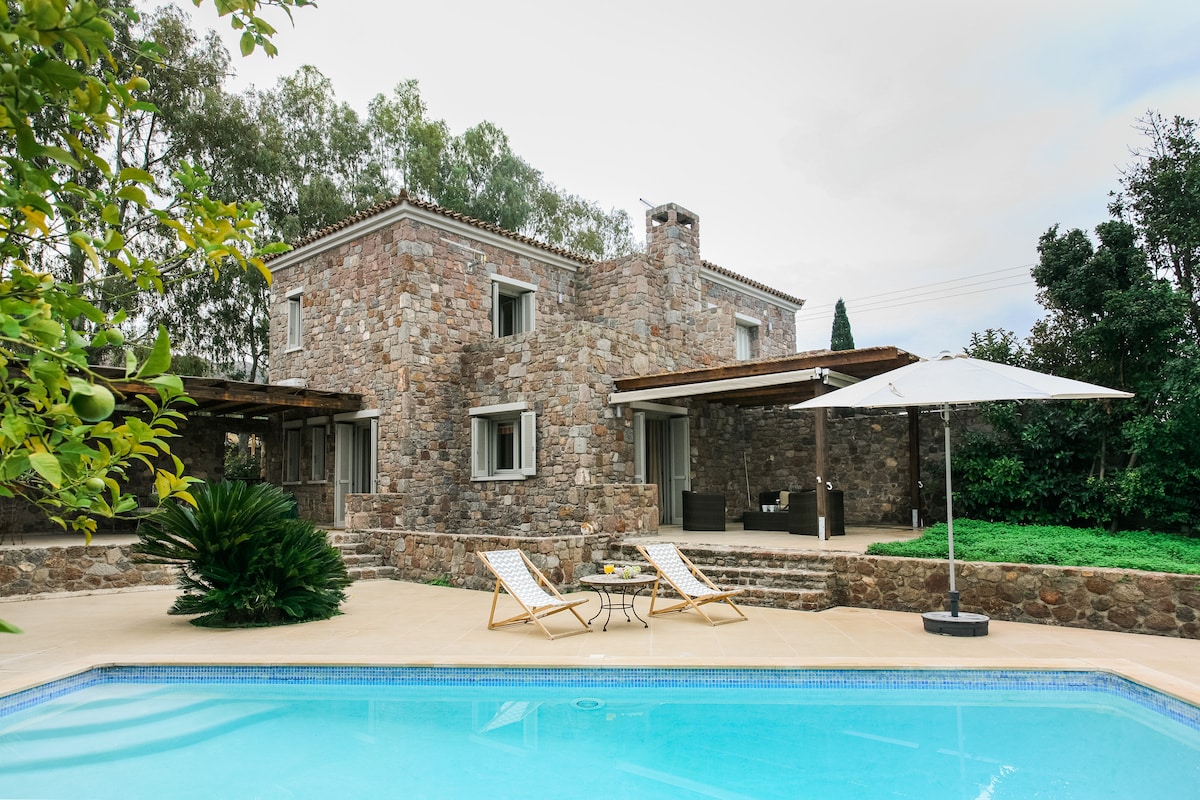 A stone-built house is set amidst lush greenery, featuring a private swimming pool. Sun loungers are positioned beside the pool, offering a comfortable spot to relax. A shaded outdoor seating area is visible, providing a tranquil space for gatherings.