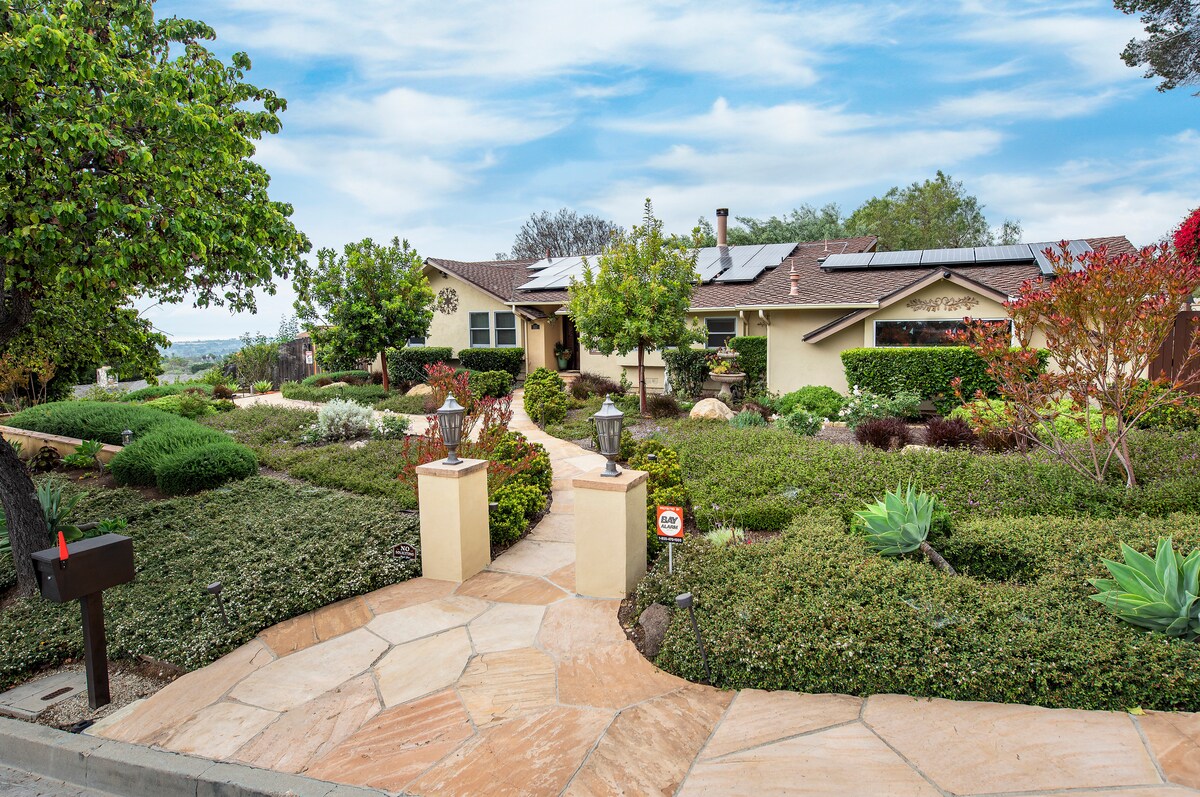 A welcoming entrance features a stone pathway lined with neatly trimmed hedges and colorful shrubs. The home is surrounded by landscaped greenery and trees, with solar panels visible on the roof. Scenic views of the surrounding hills are visible in the distance.