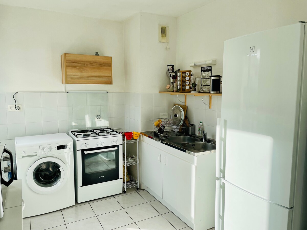 A functional kitchen is equipped with essential appliances, including a washing machine, a stove, and a refrigerator. Light tiles cover the floor, and a wooden cabinet provides storage above a stainless steel sink. Natural light brightens the uncluttered space.