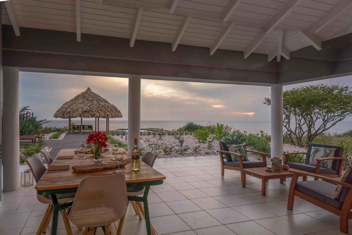 A shaded outdoor dining area is set with a wooden table adorned with fresh flowers and tableware. Secluded lounge chairs surrounding the table offer a view of a thatched-roof gazebo and the serene sea, complemented by blooming greenery in the background.
