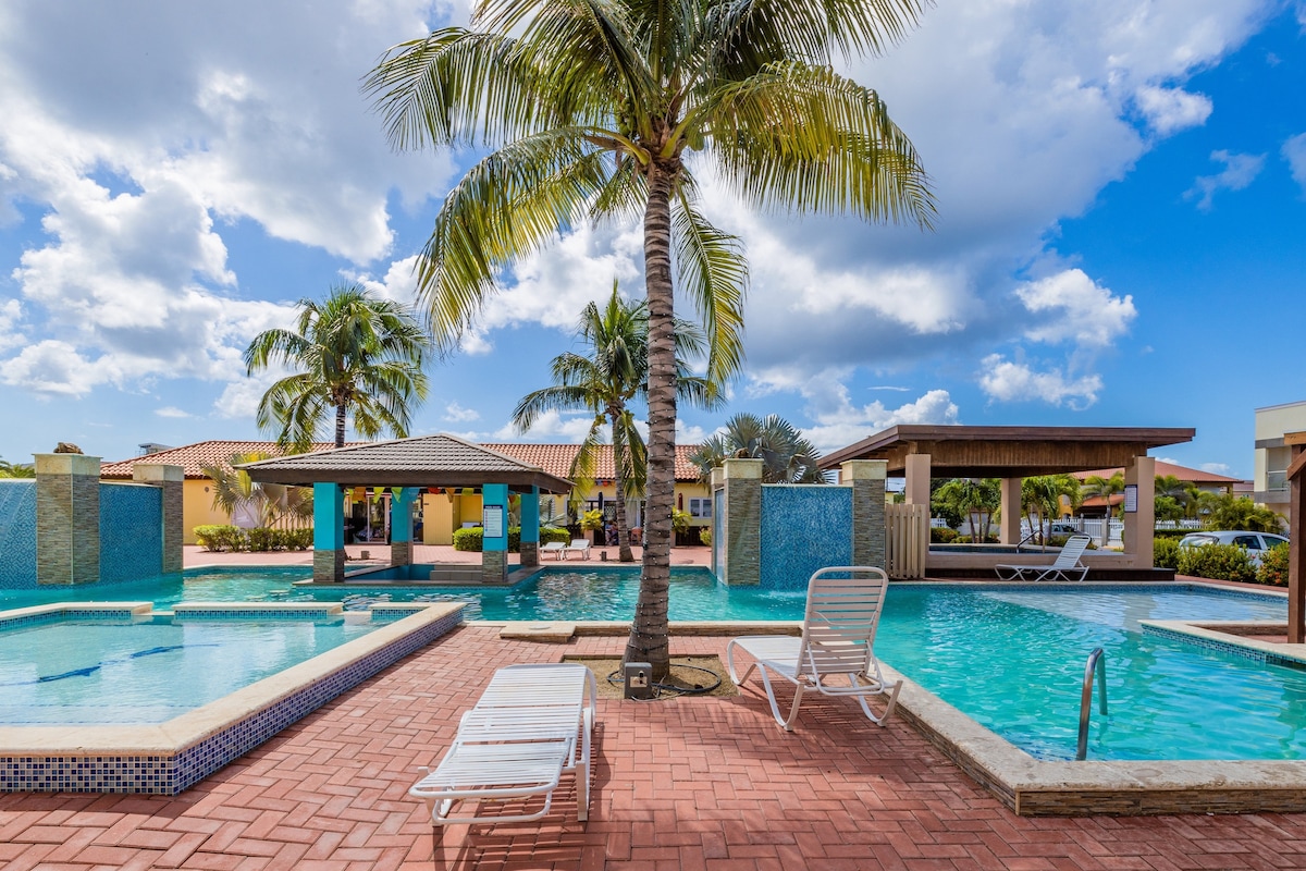 A resort-style pool area features two swimming pools surrounded by brick pathways and lush landscaping. Palm trees provide shade, while a covered seating area offers a place to relax. Clear blue skies and fluffy clouds add to the serene atmosphere of the outdoor space.