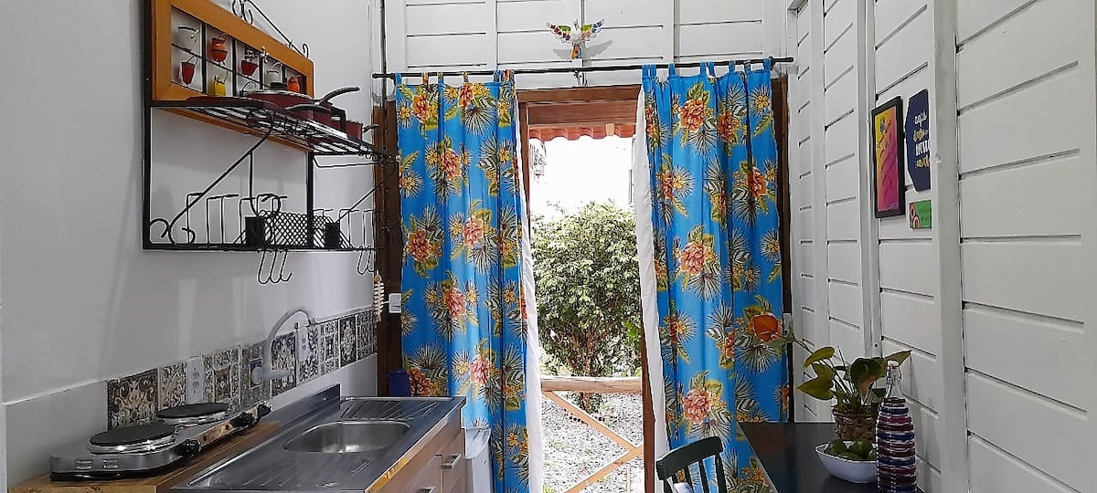 A functional kitchen area is visible, featuring a sink and stove atop a wooden counter. Brightly patterned curtains frame an open doorway leading to a garden view. Decorative items and plants add a touch of color and life to the space.