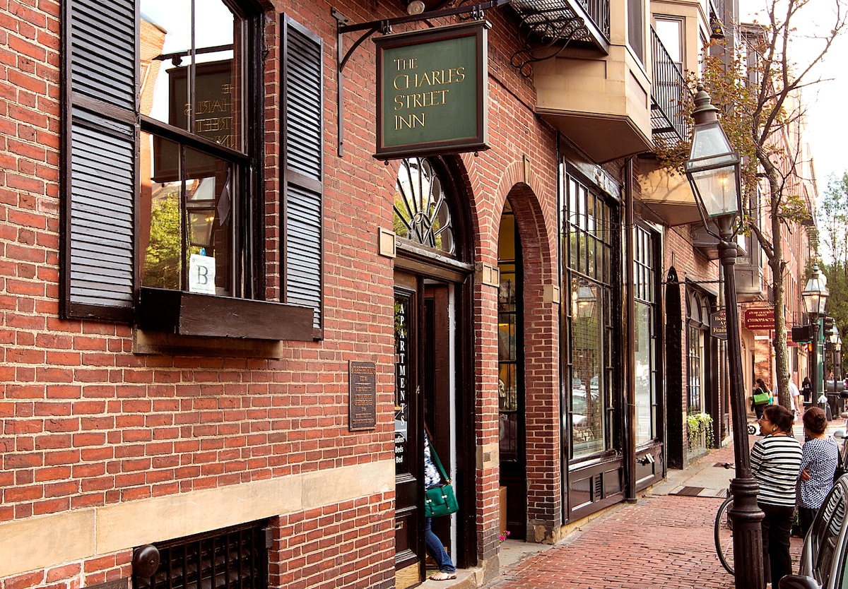 The image displays a charming brick building, showcasing large windows adorned with decorative black shutters. A sign reading 'The Charles Street Inn' hangs above the entrance, while cobblestone streets are visible in front, lined with outdoor seating and greenery, reflecting the historic ambiance of Beacon Hill.