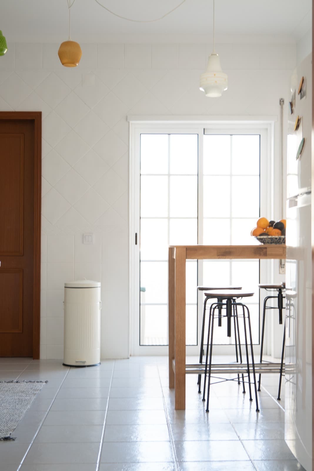 The kitchen features a modern wooden dining table accompanied by three sleek bar stools. A bowl of fresh fruit rests on the table. Large sliding glass doors allow natural light to fill the space, and a white trash bin is positioned nearby.