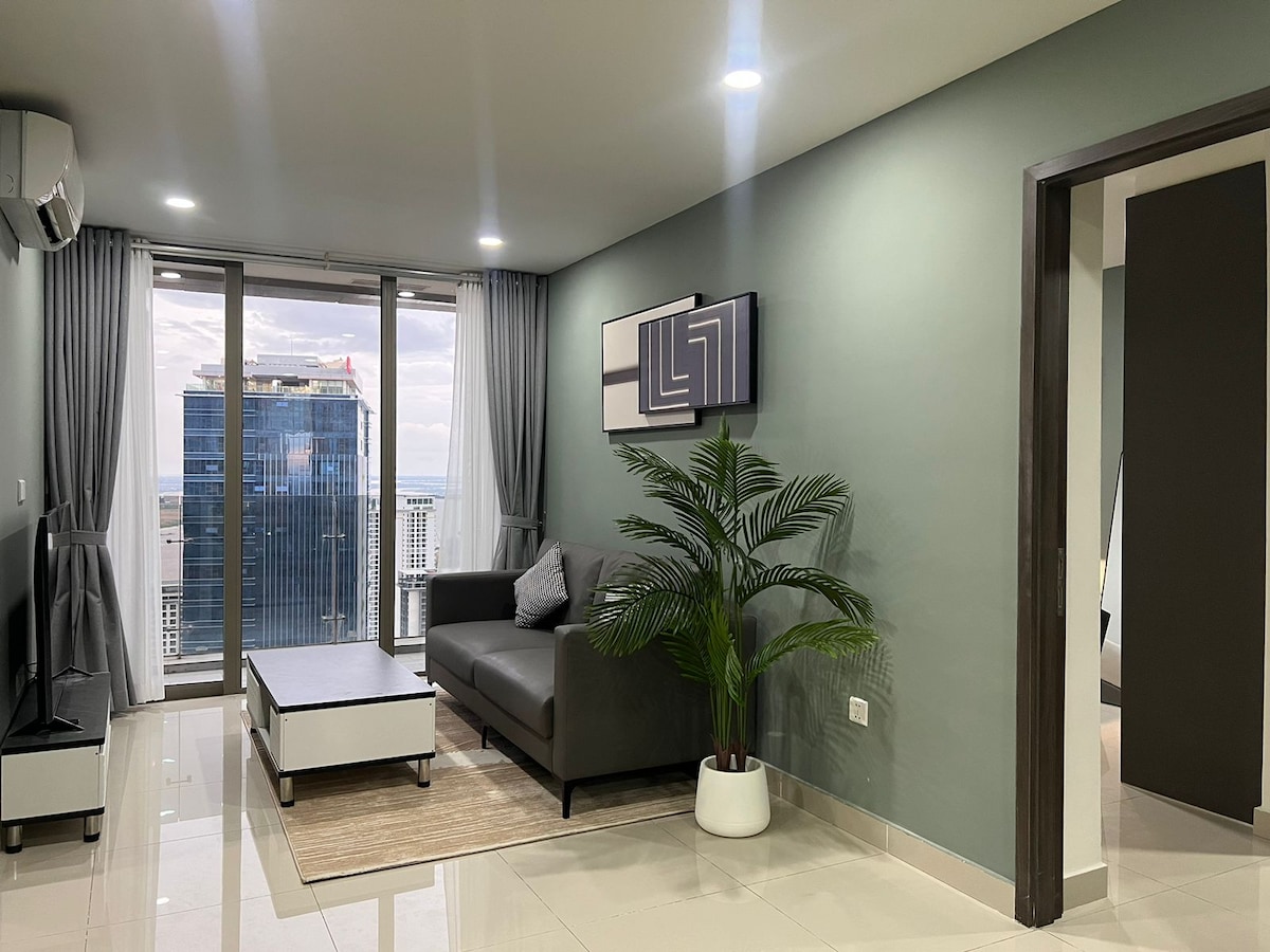 A modern living area includes a comfortable sofa, a coffee table, and a decorative plant. Large windows provide views of the city landscape. Neutral wall colors and sleek furnishings contribute to a relaxed atmosphere.