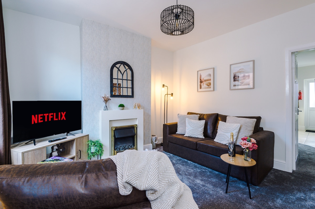 A modern living area is presented with a cozy sofa set, complemented by decorative pillows. A sleek smart TV displays the Netflix logo, while soft lighting is provided by a standing lamp. Neutral tones and a decorative fireplace enhance the inviting atmosphere.