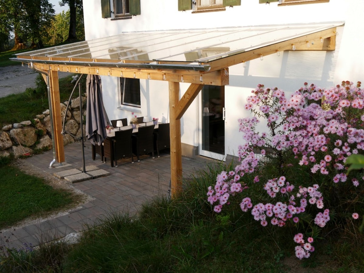 A covered outdoor terrace features a wooden structure with a clear roof, providing shade and shelter. A dining table set for six is placed on a paved surface. Surrounding the terrace, flowering plants add color to the landscape.