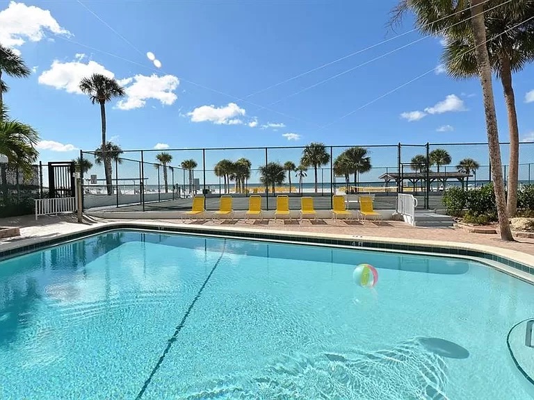 A heated pool is featured in the foreground, showcasing clear blue water. Yellow loungers are arranged nearby, with palm trees visible beyond a fence. The background presents a serene beach and bright blue sky dotted with fluffy clouds.