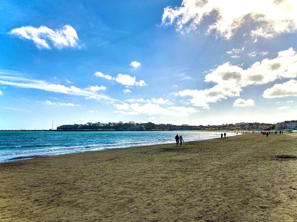 A broad sandy beach stretches along the water’s edge, where gentle waves meet the shore. The sky is partly cloudy, with sunlight illuminating the scene. People can be seen walking leisurely along the beach, with a distant shoreline visible in the background.