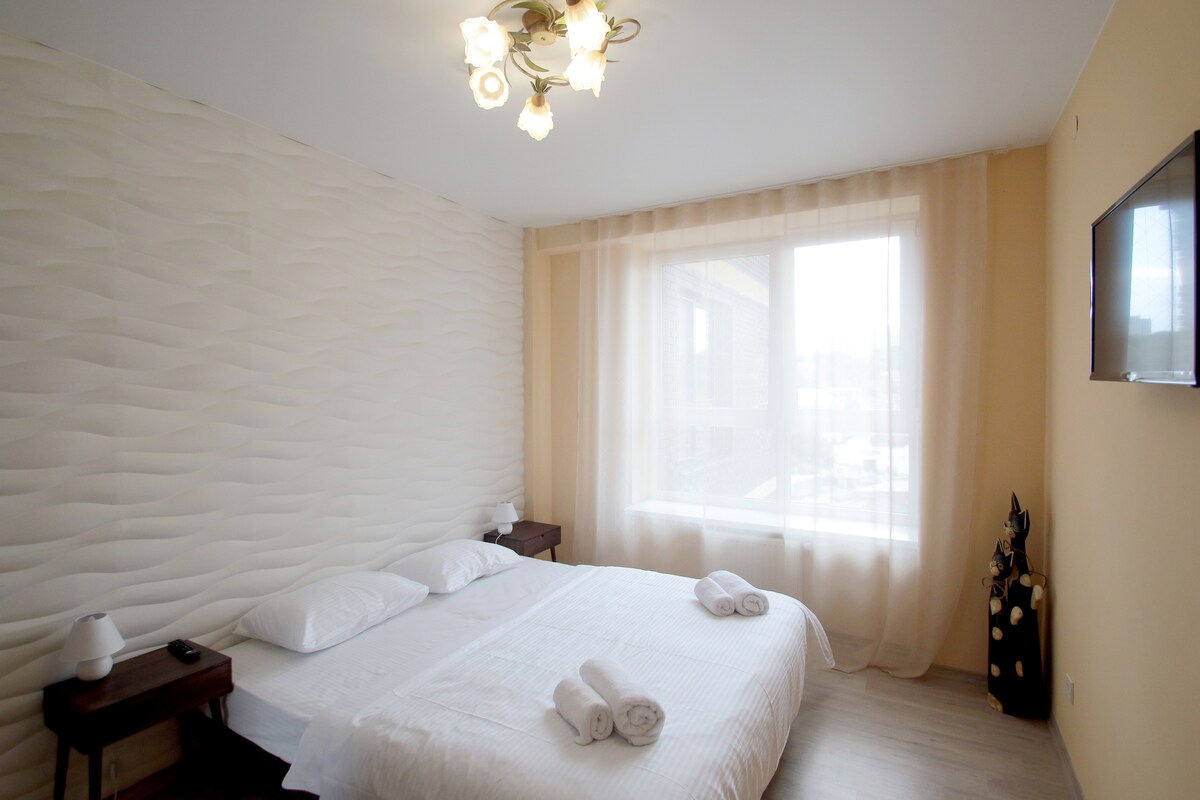 A spacious room features a large window dressed with sheer curtains, allowing natural light to enter. A neatly made bed is complemented by two bedside tables with lamps on either side. Decorative elements, including a sculpture, are present in the corner.