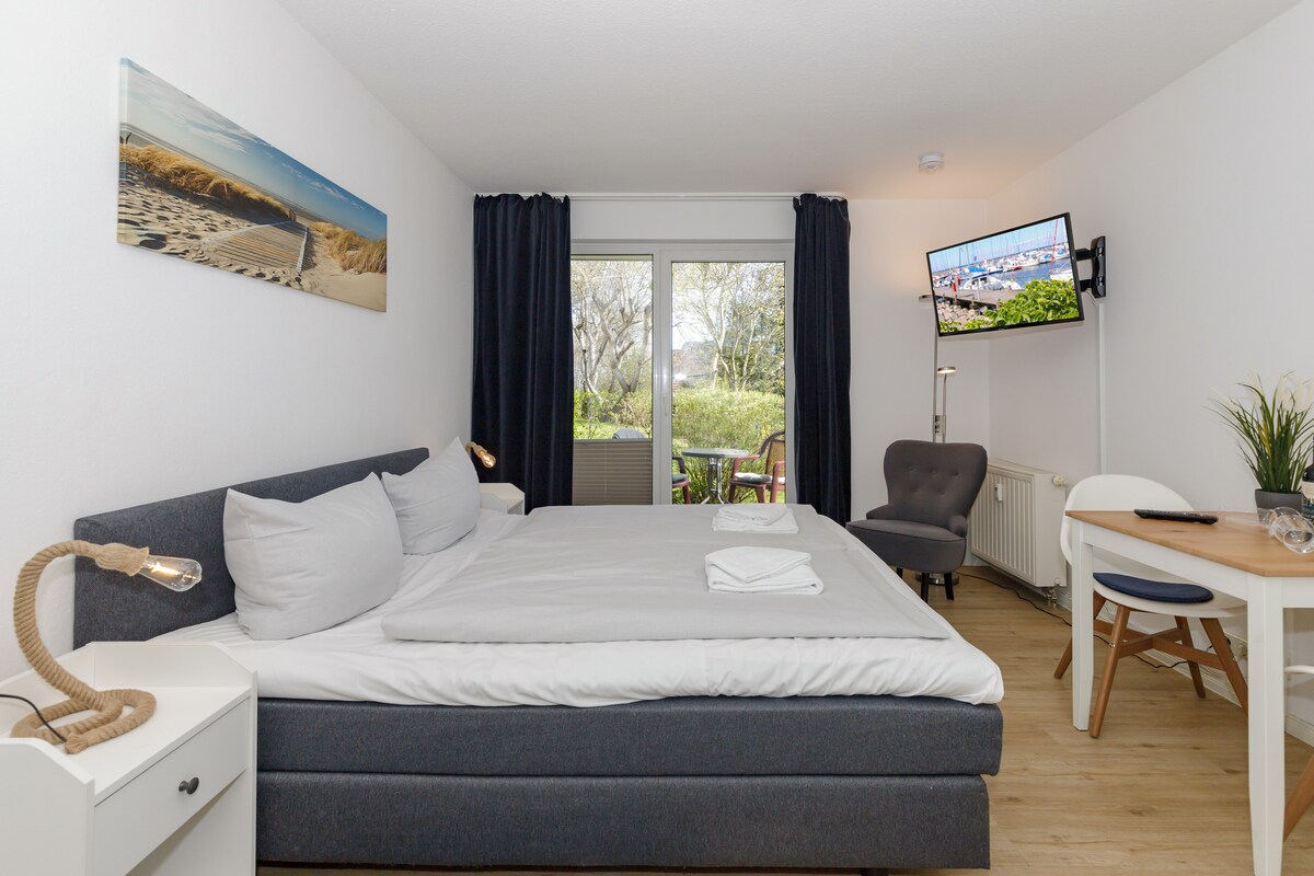 A light and airy living space features a spacious bed with crisp linens, accompanied by decorative accents. A comfortable chair is positioned nearby, and a small dining table with two chairs is visible. A television is mounted on the wall, and large windows open to a tranquil terrace.