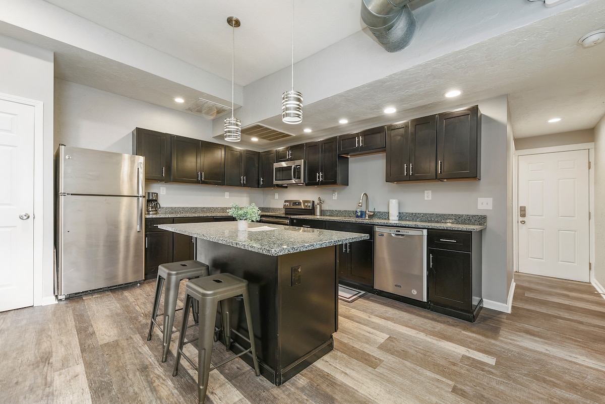 A modern kitchen features dark cabinetry alongside stainless steel appliances, complemented by a spacious granite countertop. Three metal stools are positioned at the island, and ample lighting illuminates the area, including ceiling fixtures and under-cabinet lights. The layout encourages convenience and ease for cooking.