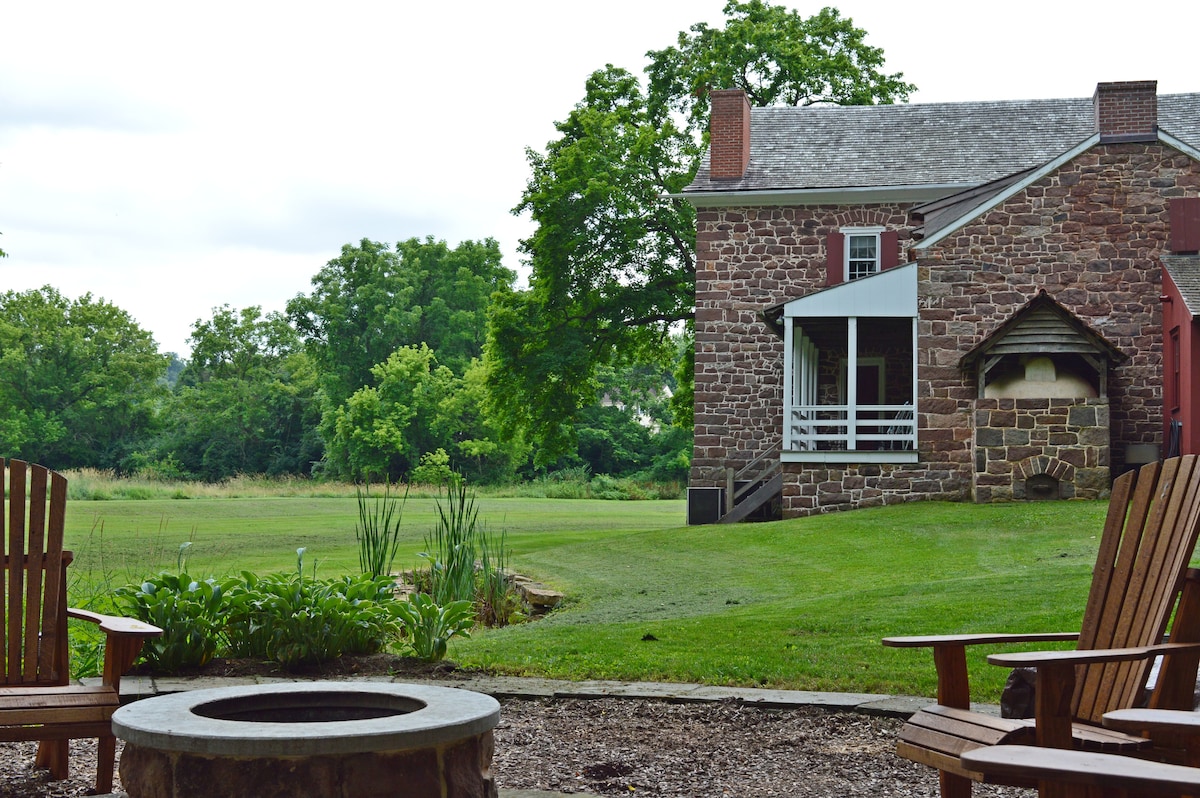 A peaceful outdoor space features a fire pit surrounded by wooden chairs. The historic stone house is visible in the background, complemented by lush green grass and trees, creating a serene atmosphere for relaxation.
