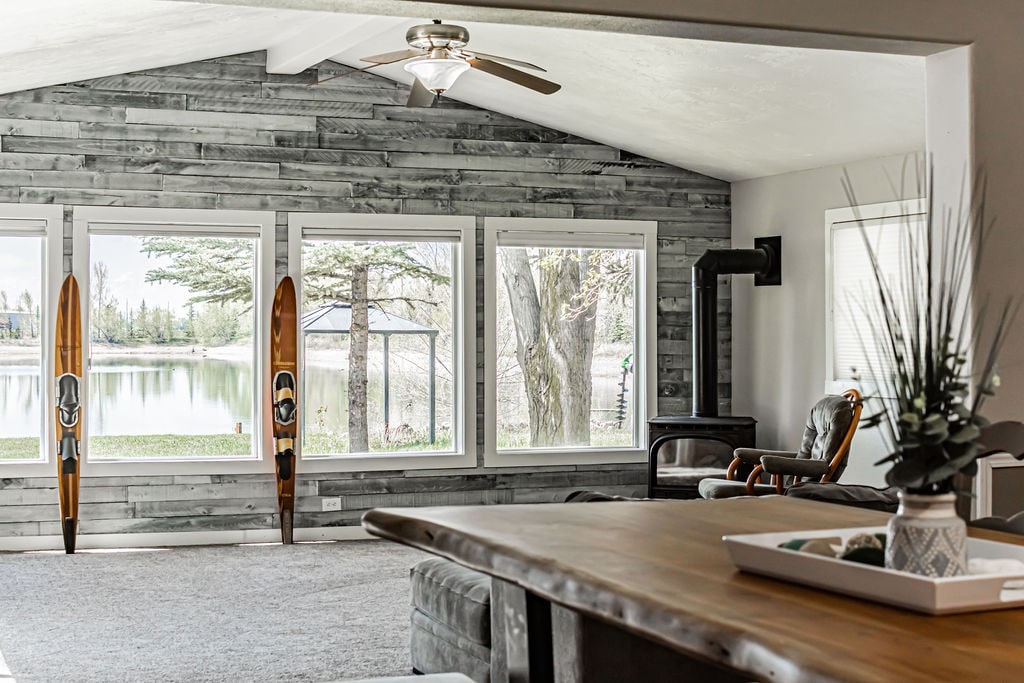 A bright living area is showcased, featuring large windows that frame views of the lake outside. Two wooden paddleboards are displayed near the windows, and a comfortable seating area includes a leather chair and a wooden table. Light-colored walls and a ceiling fan add to the space.