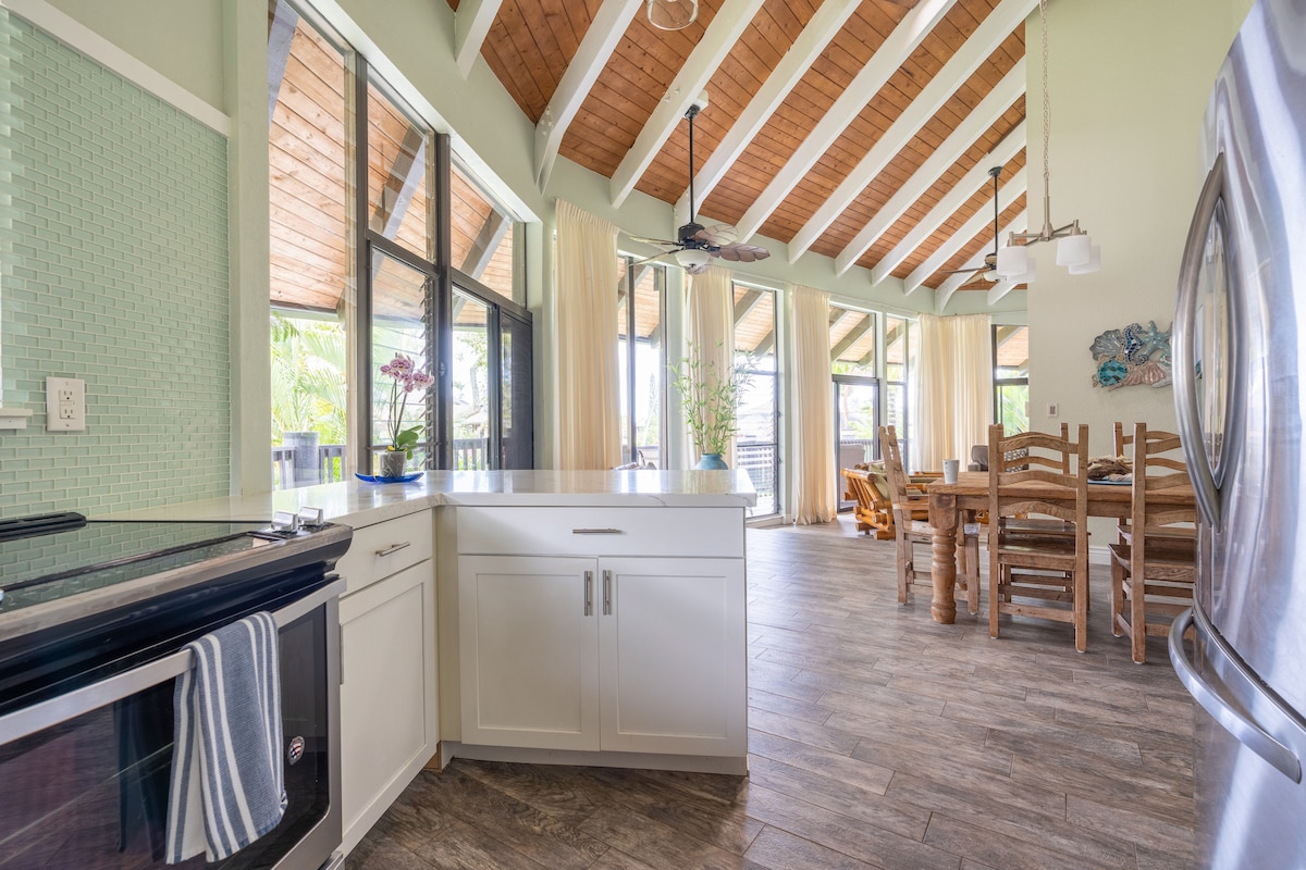 An open kitchen area is highlighted by a combination of white cabinetry and natural wood accents. Large windows allow ample natural light, illuminating the dining table and a set of wooden chairs nearby. The vaulted ceiling features exposed wooden beams, enhancing the spacious feel.