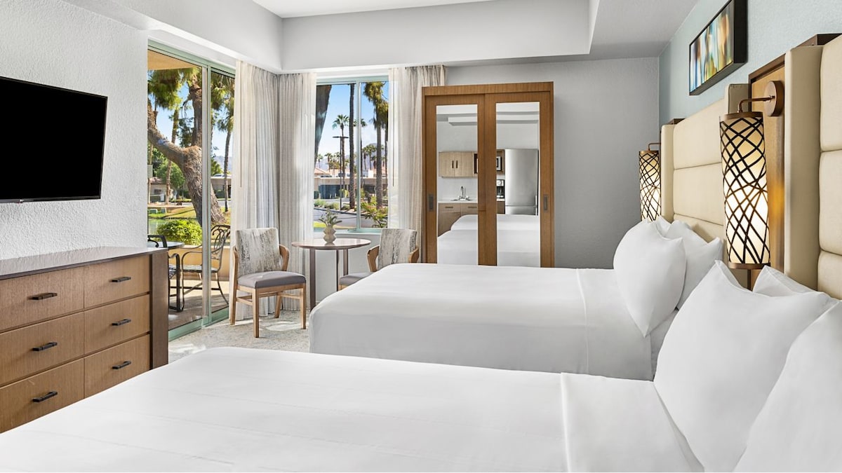 A spacious guest room is furnished with two queen beds, side tables, and a dresser. Large glass doors open to a view of the outdoors, featuring palm trees. A small dining table with two chairs is situated near the windows, allowing for natural light.