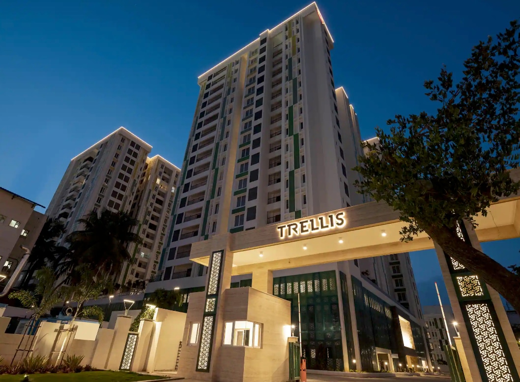 A modern high-rise building is illuminated at dusk, highlighting its sleek architecture. The entrance is marked by a prominent sign reading 'TRELLIS', with lush greenery framing the entrance and adaptive lighting enhancing the contemporary facade.