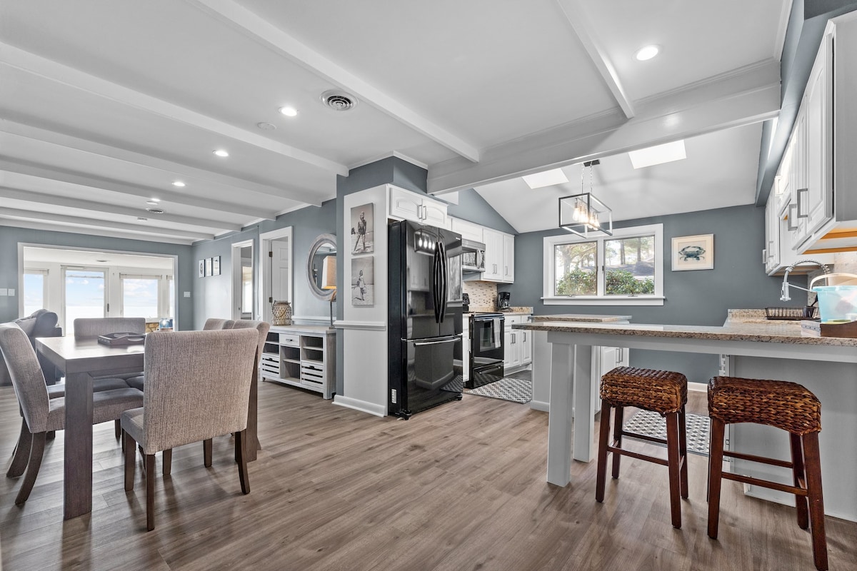 A stylish kitchen and dining area are displayed, featuring sleek appliances and light wood flooring. Natural light floods through the windows, illuminating the dining table and chairs, as well as the breakfast bar with two stools. Soft blue walls create a serene atmosphere.