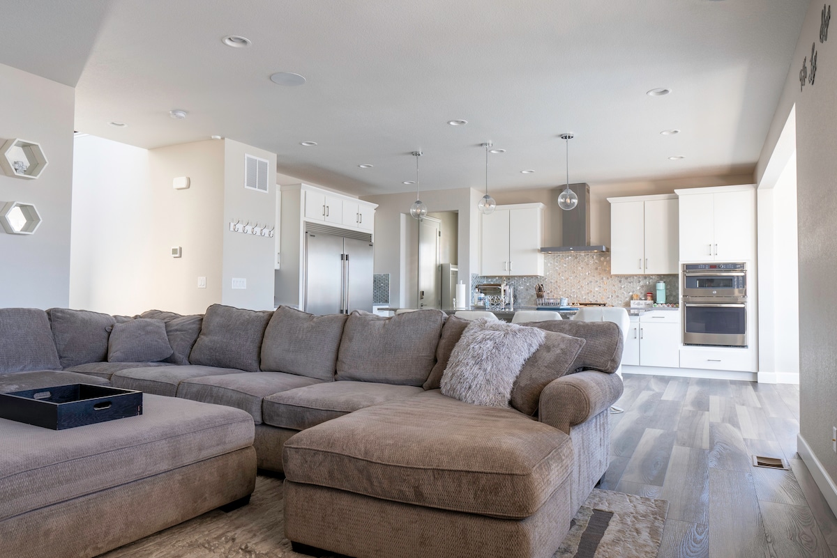 A spacious living area is filled with natural light from large windows. A comfortable sectional sofa occupies the foreground, while the modern kitchen is visible in the background, featuring white cabinetry and stainless steel appliances. Seamless flow is created between the living and dining spaces.