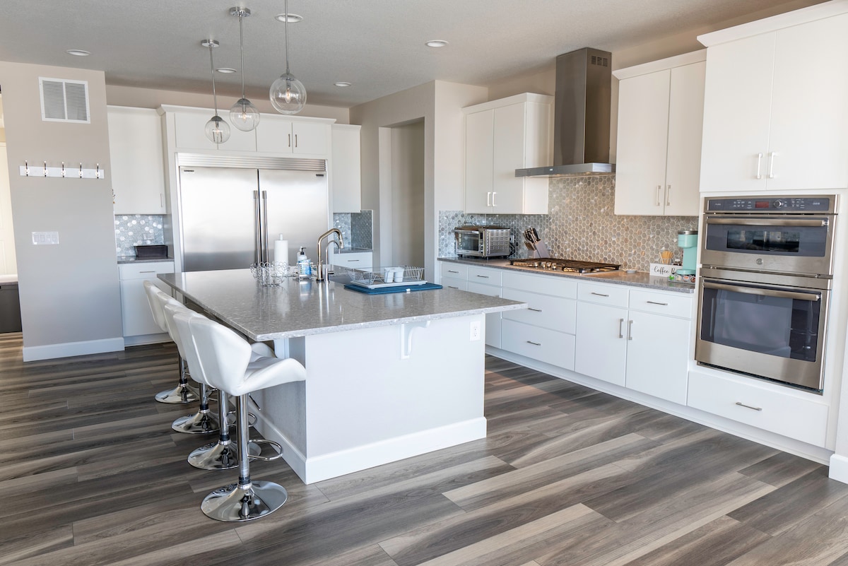 A modern kitchen is highlighted by sleek white cabinetry and stainless steel appliances. A large island with seating is centered in the space. Warm wood-like flooring complements the contemporary design. Pendant lights hang above the island, creating a well-lit atmosphere.
