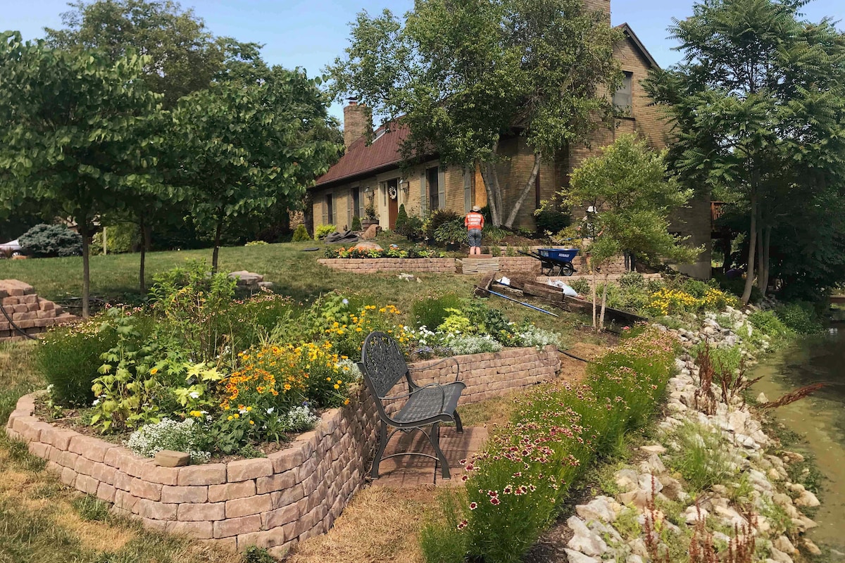 A well-maintained garden with a variety of colorful flowers and greenery is featured in the foreground, bordered by a stone wall. Shade is provided by several trees, while the serene Tara Lake is visible alongside the lush landscaping.