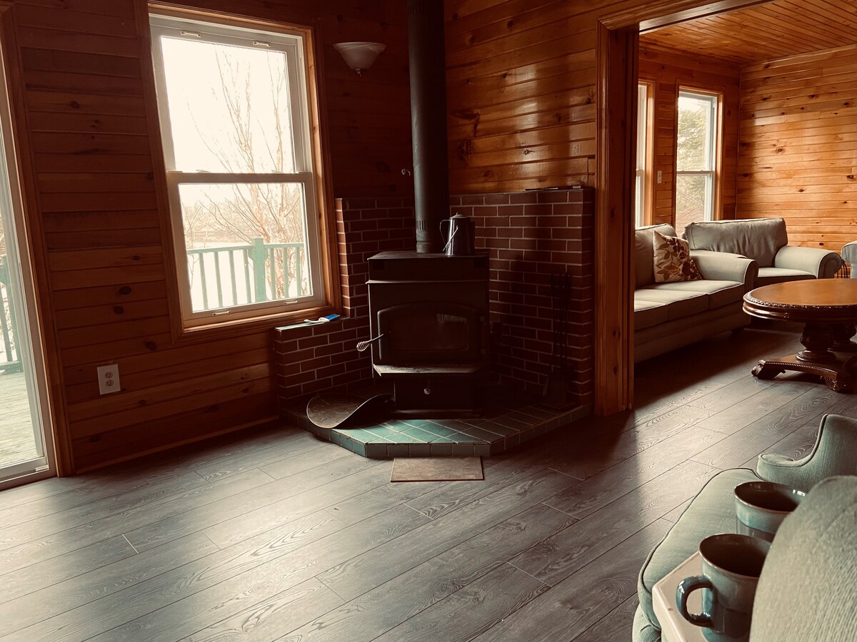 A cozy interior features warm wooden walls and a wood-burning stove set on a slate base. A comfortable seating area is visible with a soft sofa and a round table nearby. Natural light enters through multiple windows, enhancing the inviting space.