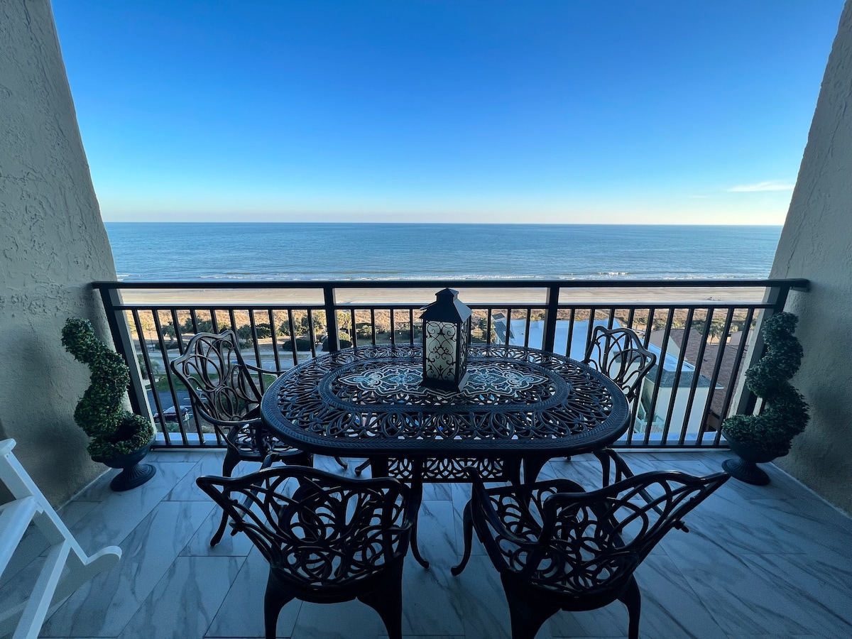 A spacious balcony features an ornate metal table and four matching chairs, offering direct ocean views. A large lantern stands at the center of the table, with the tranquil ocean and beach in the background. The scene is framed by decorative plants on either side.